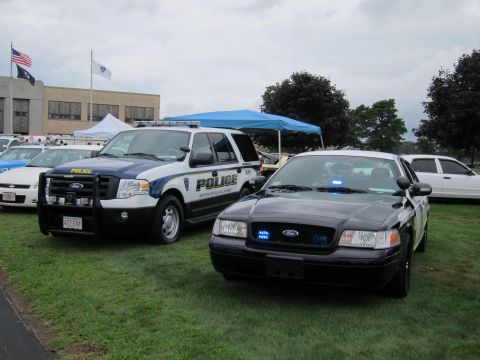1991 Ford LTD Crown Victoria - black Ford Crown Victoria police cruiser and a white police SUV parked on grass at an outdoor event with flags and a tent.