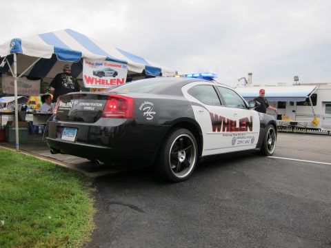 1991 Ford LTD Crown Victoria - Rear three-quarter view of a black-and-white Dodge Charger demonstration vehicle wrapped with Whelen branding beside a vendor tent.