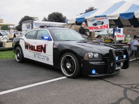 1991 Ford LTD Crown Victoria - Front three-quarter view of a black-and-white Dodge Charger with push bumper and blue lights, marked Whelen, parked near vendor tents.