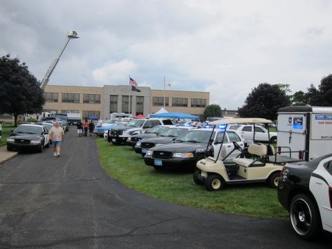 1991 Ford LTD Crown Victoria - line of police cruisers, a golf cart and trailers displayed on a lawn at an outdoor event with a raised fire-truck ladder behind.