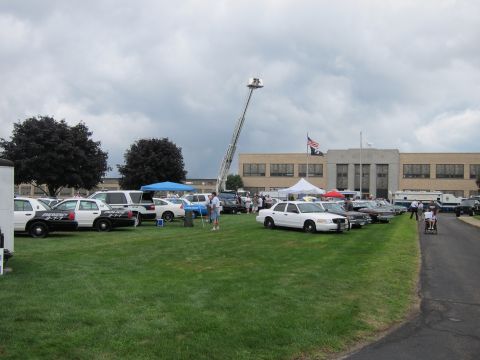 1991 Ford LTD Crown Victoria - Police cruisers and white sedans parked on a grassy field under a cloudy sky, with a fire department aerial ladder extended in back.