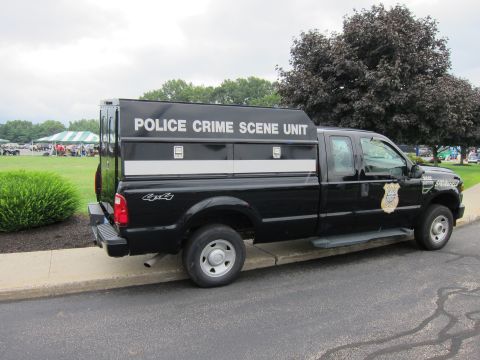 1991 Ford LTD Crown Victoria - Rear three-quarter view of a black 4x4 pickup with a Police Crime Scene Unit bed cap parked at the curb near event tents.