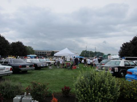 1991 Ford LTD Crown Victoria - Rows of vintage police cars line a grassy car-show field with a white event tent and building under an overcast sky.