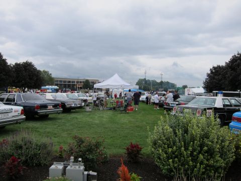 1991 Ford LTD Crown Victoria - grassy show field crowded with classic police cruisers, attendees, and a white tent beneath an overcast sky.
