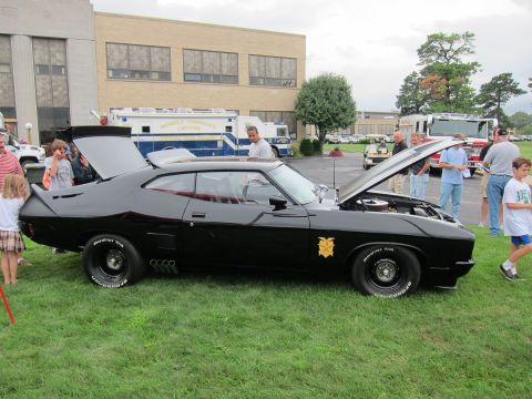 1991 Ford LTD Crown Victoria - black customized muscle car with a tall rear spoiler and open trunk on display, spectators gathered around on a grassy lot.