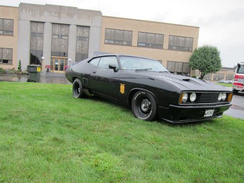 1991 Ford LTD Crown Victoria - black fastback muscle car with a hood scoop, gold door emblem and 'MFP' plate sits on grass before an art-deco building.