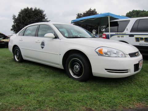 1991 Ford LTD Crown Victoria - white Chevrolet Impala police-style sedan with steel wheels and a small door decal parked on grass beside a canopy tent.