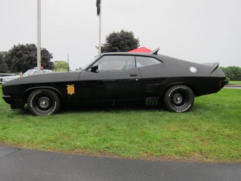 1991 Ford LTD Crown Victoria - Side profile of a black customized fastback muscle car showing side exhaust pipes, gold door emblem and white-letter tires.