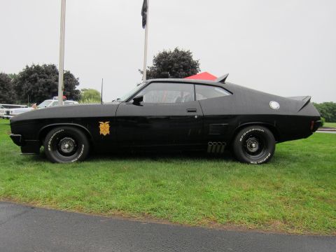 1991 Ford LTD Crown Victoria - Profile of a black customized fastback muscle car with side pipes and gold door emblem parked on grass before a tree line.