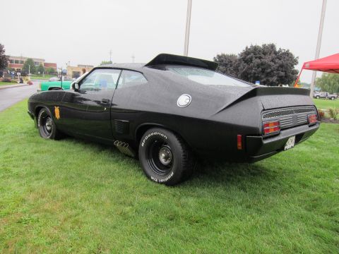 1991 Ford LTD Crown Victoria - Rear view of a black customized fastback muscle car showing side exhaust pipes, gold emblem and white-letter tires on grass.
