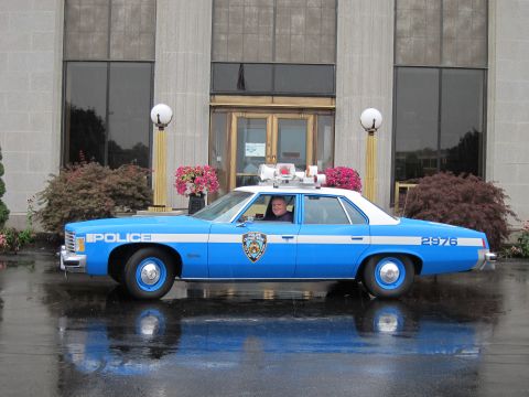1991 Ford LTD Crown Victoria - Side view of a blue 'POLICE' sedan numbered 2976 with roof lightbar parked before a glass-and-brass building entrance.
