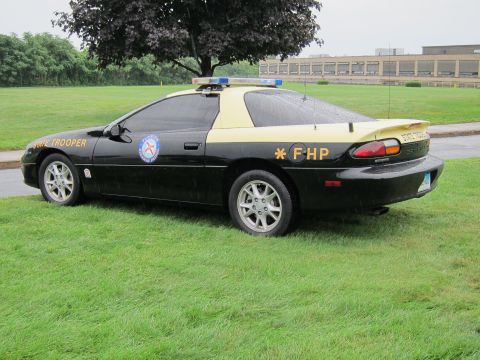 1991 Ford LTD Crown Victoria - Rear three-quarter view of a black-and-tan Chevrolet Camaro 'FHP State Trooper' cruiser with roof lightbar on a grassy field.