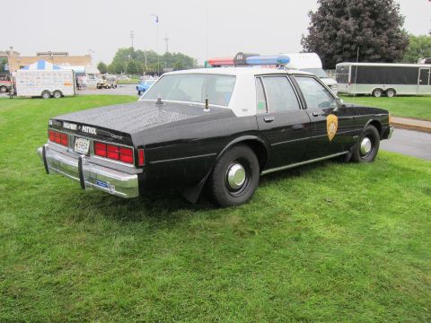 1991 Ford LTD Crown Victoria - Rear quarter view of a black police cruiser marked Highway Patrol with a roof light bar, parked on grass near trailers.