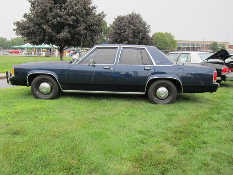 1991 Ford LTD Crown Victoria - dark blue four-door sedan with steel wheels and full hubcaps shown in side profile on a grass lawn at a car show.