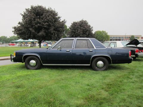 1991 Ford LTD Crown Victoria - Driver-side profile of a dark blue full-size four-door sedan parked on grass with trees and buildings behind.