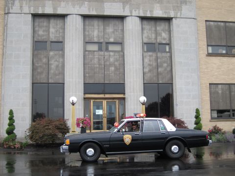 1991 Ford LTD Crown Victoria - black-and-white police sedan with roof lights parked on wet pavement in front of a stone civic building entrance.
