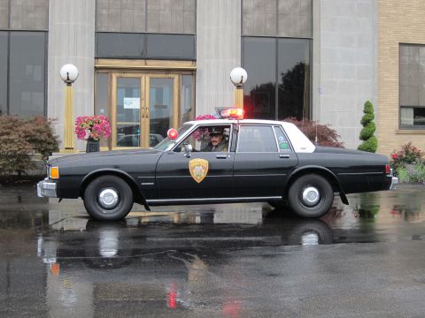 1991 Ford LTD Crown Victoria - Side view of a black-and-white police cruiser with activated red roof lights on rain-soaked pavement, a person inside.