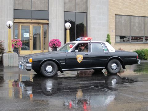 1991 Ford LTD Crown Victoria - black-and-white police sedan with glowing red roof lights and a driver, reflected on the wet pavement before a building.