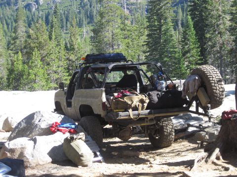 1989 Ford Bronco - A heavily modified off-road vehicle is parked in a wooded, rocky area, laden with supplies for an outdoor excursion.