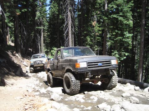1989 Ford Bronco - Two off-road vehicles navigate a challenging, rocky trail through a dense forest.