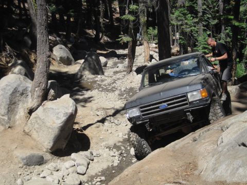 1989 Ford Bronco - A 1989 Ford Bronco tackles a challenging, rocky off-road path with a person standing by the driver's door.