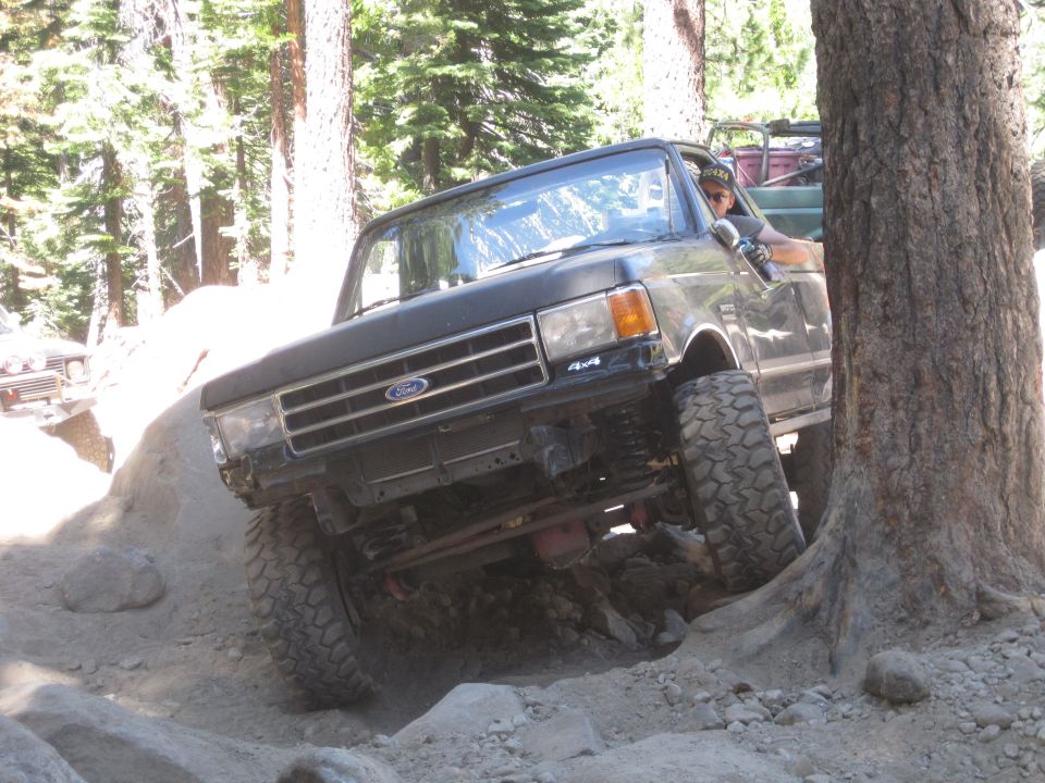 Ford F-Series truck navigating rocky terrain near a tree. Another vehicle is visible in the background