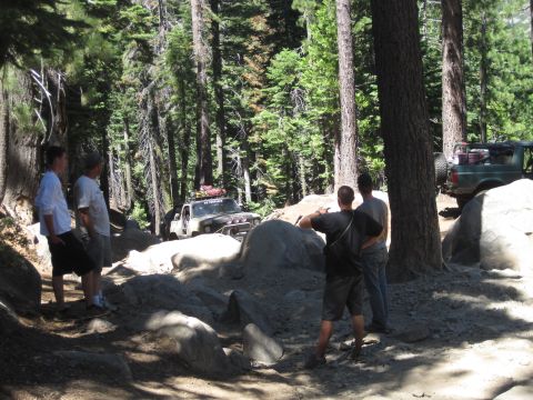 1989 Ford Bronco - Several off-road vehicles navigate a challenging, rocky trail through a dense forest, with onlookers observing the action.