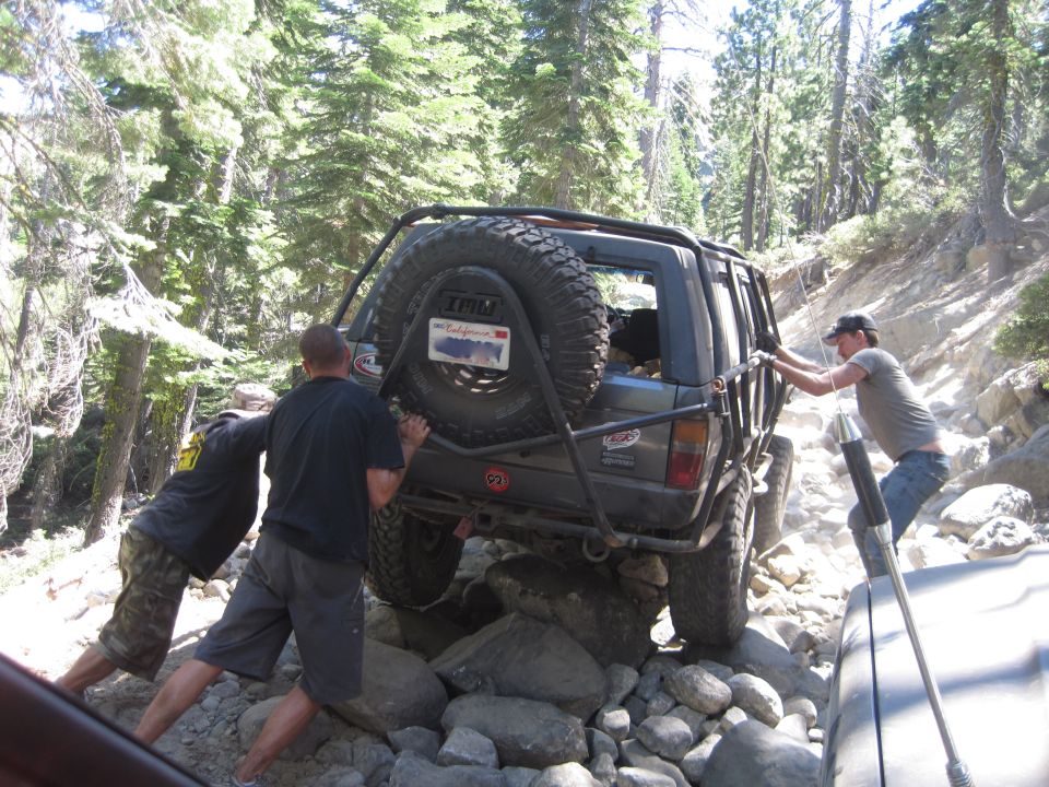 Off-road SUV stuck on rocks, people pushing it. Another vehicle visible