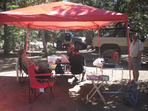 1989 Ford Bronco - People relax and cook outdoors near two off-road vehicles parked in a wooded area.