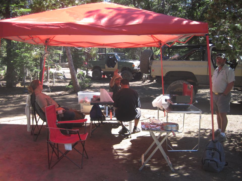 Two off-road vehicles, one green and one tan, parked in a wooded area under a red canopy with people sitting and standing