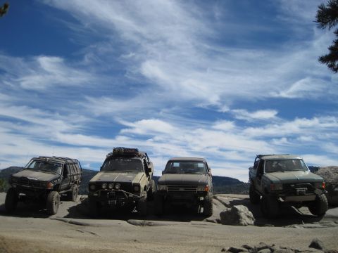1989 Ford Bronco - Multiple modified off-road vehicles are parked on a rocky landscape with a dramatic sky overhead.