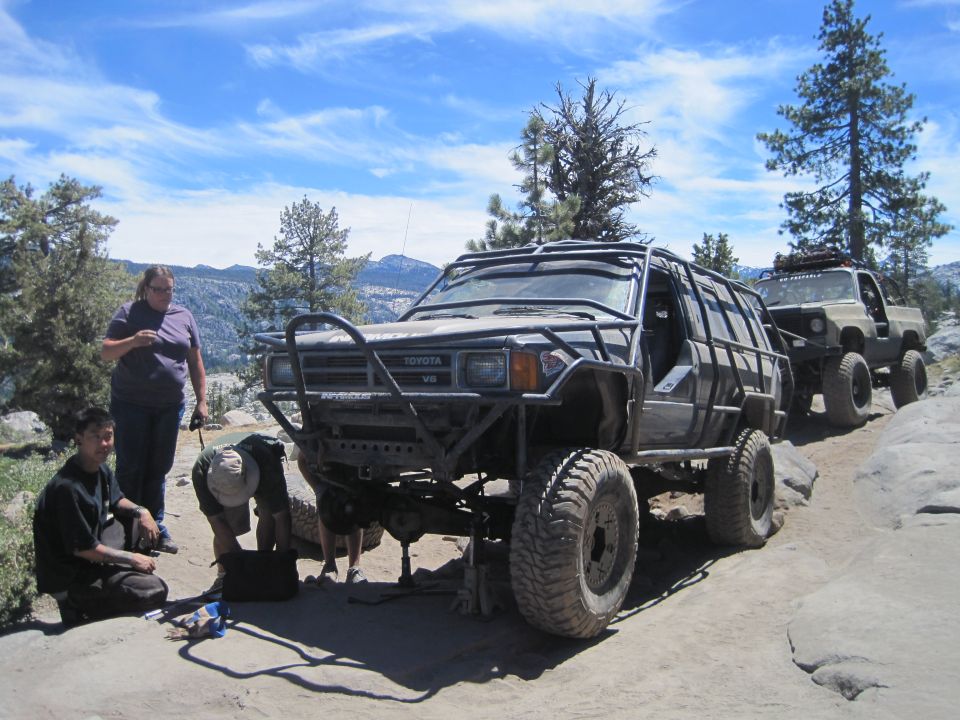 Toyota 4Runner with custom roll cage and bumper, people working on it, another truck behind it on a dirt trail