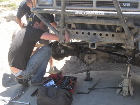 1989 Ford Bronco - Mechanics work on the front axle of an off-road vehicle, which has a V6 engine and "Infamous" branding on the grille guard.