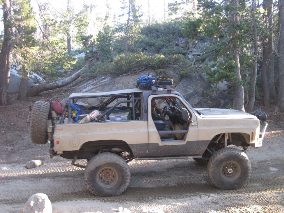 Modified SUV with external roll cage, spare tire on rear, and roof rack, parked on a dirt road in a wooded area