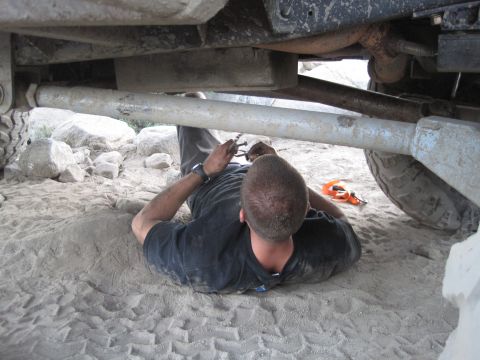 1989 Ford Bronco - A person lies on the ground working on the undercarriage of a vehicle, with rocks and sand visible.