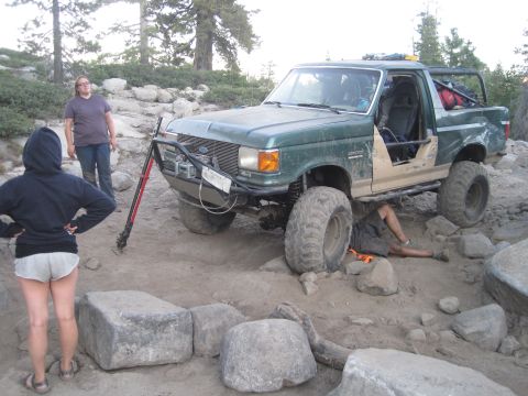 1989 Ford Bronco - A green Ford Bronco is being worked on by a person underneath it, with two other people standing nearby.
