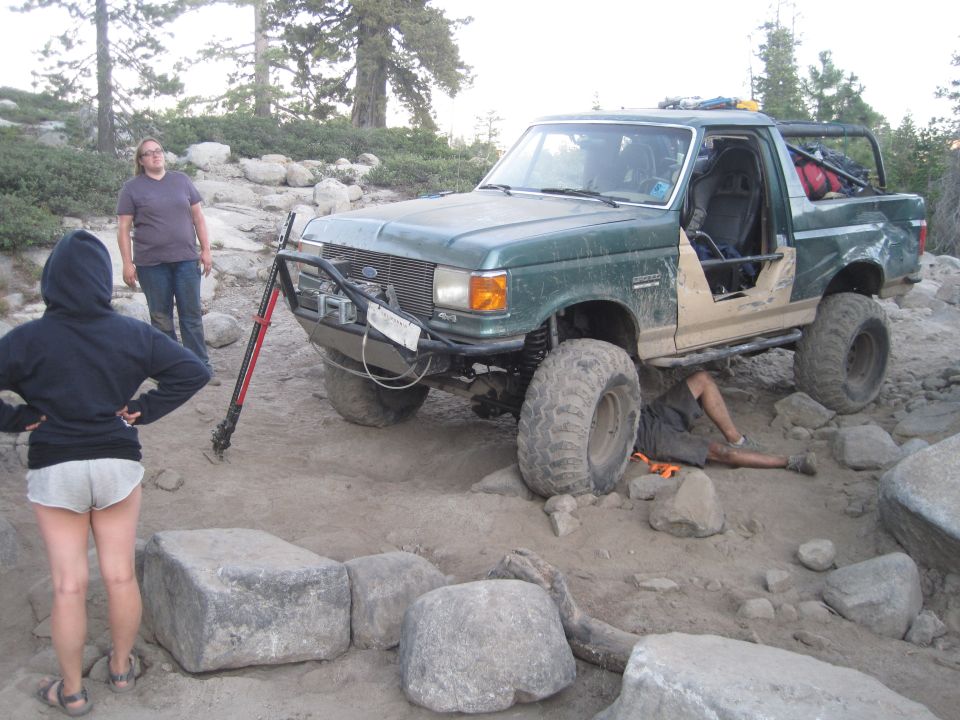 Green Ford Bronco with off-road modifications, driver's door removed, person working underneath