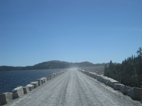 1989 Ford Bronco - A dusty gravel road stretches towards a dam, bordered by water and trees.