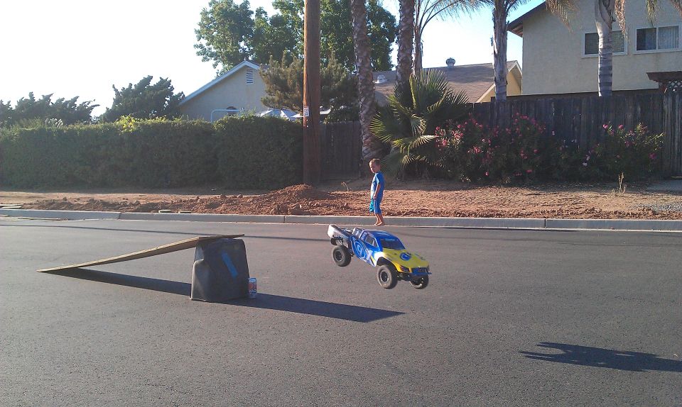 Yellow and blue remote control monster truck airborne over asphalt road, child watches