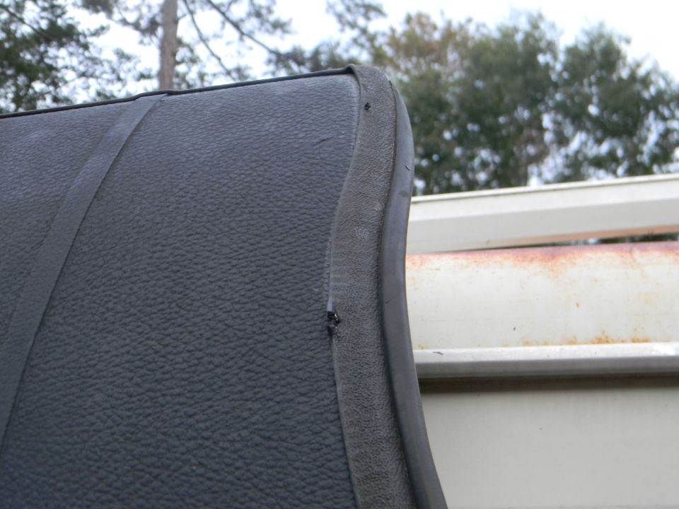 Close-up of black textured upholstery with a seam and a rubber trim piece. A white structure with rust is in the background