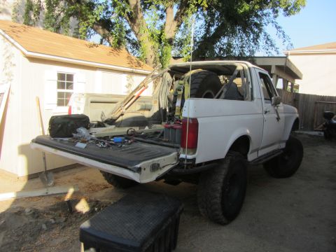 1990 Ford Bronco - A white Ford Bronco with a lifted suspension and large tires is parked in a yard with its tailgate open.