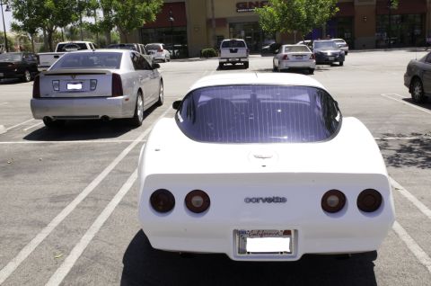 2006 Cadillac STS - A white Corvette is parked in a lot, showing its rear end and 'Corvette' badging. A silver Cadillac is parked nearby.
