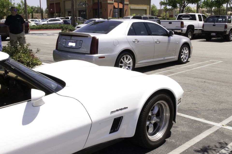White sports car in foreground, silver Cadillac sedan behind it in parking lot. Pickup trucks visible in background