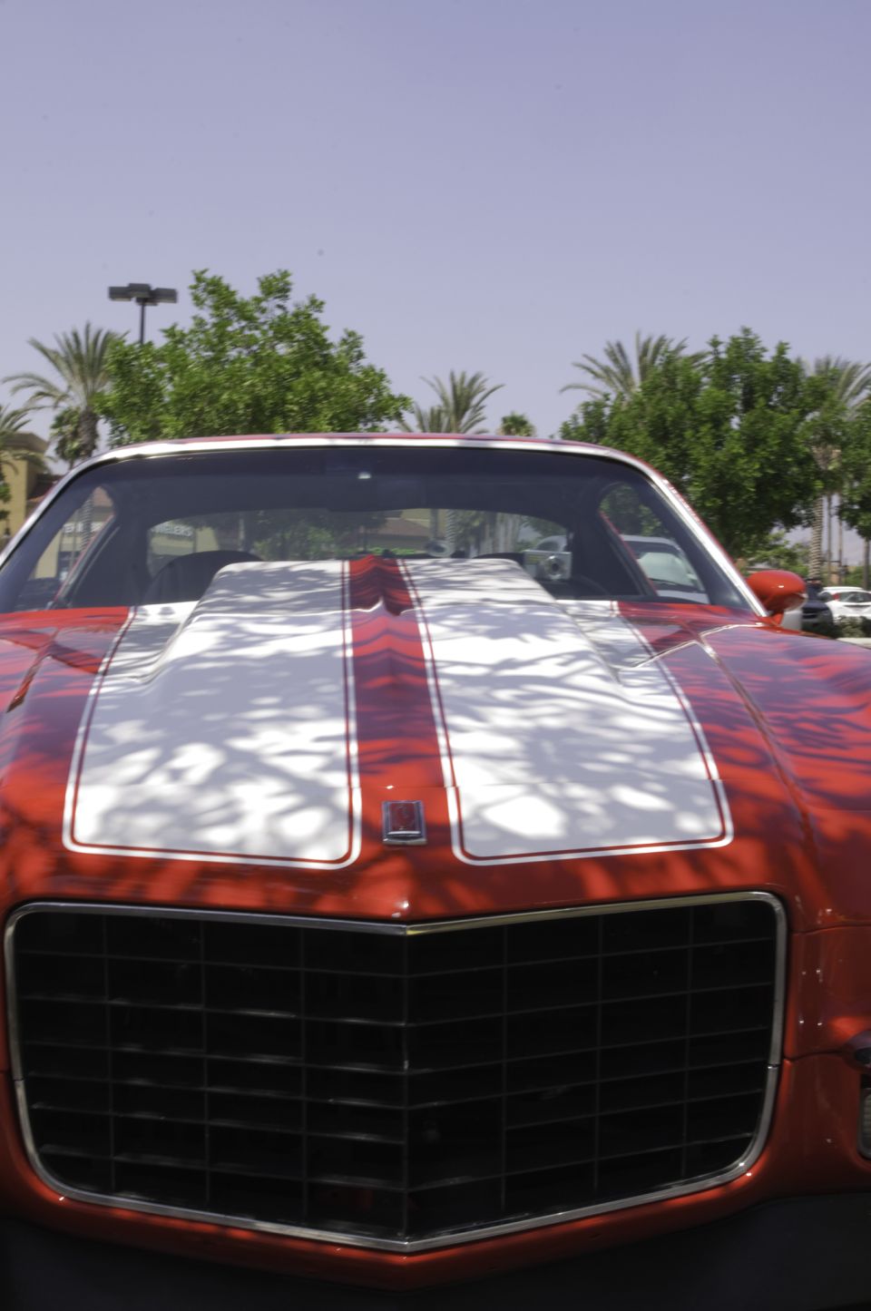 Front view of a red muscle car with a large white hood scoop and racing stripes. A small badge is on the hood