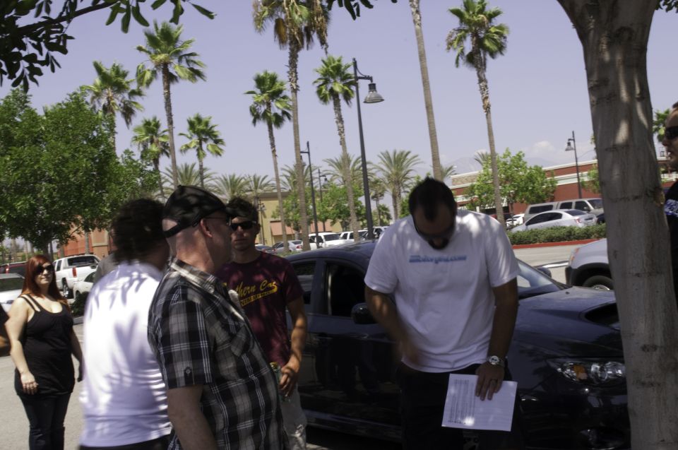 Group of people near a dark car, with palm trees and buildings in the background. A watermark says SUPERMOTORS