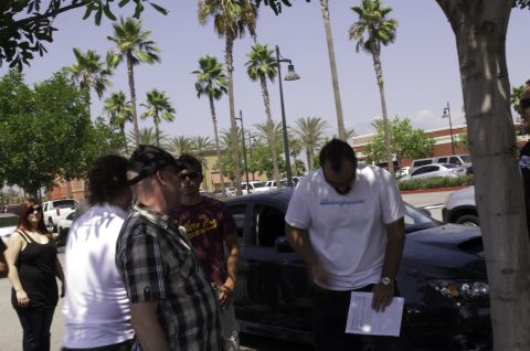 2006 Cadillac STS - Several individuals are gathered around a dark sedan in a parking lot with palm trees overhead.
