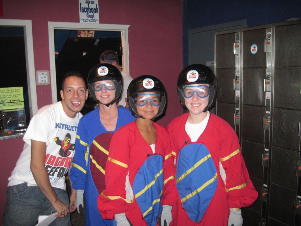 Four people in skydiving suits and helmets pose for a photo, with lockers in the background