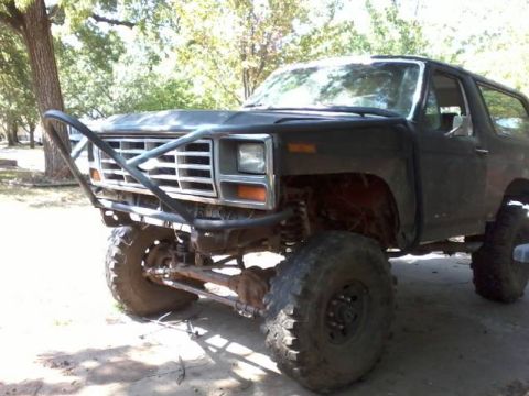 1986 Ford Bronco - Lifted 1980s Ford Bronco in flat dark green/primer, photographed from the front three-quarter under trees on a dirt lot. It runs a tubular…