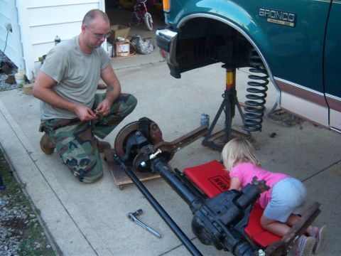 1996 Ford Bronco - A man works on the suspension of a 1996 Ford Bronco while a child watches.