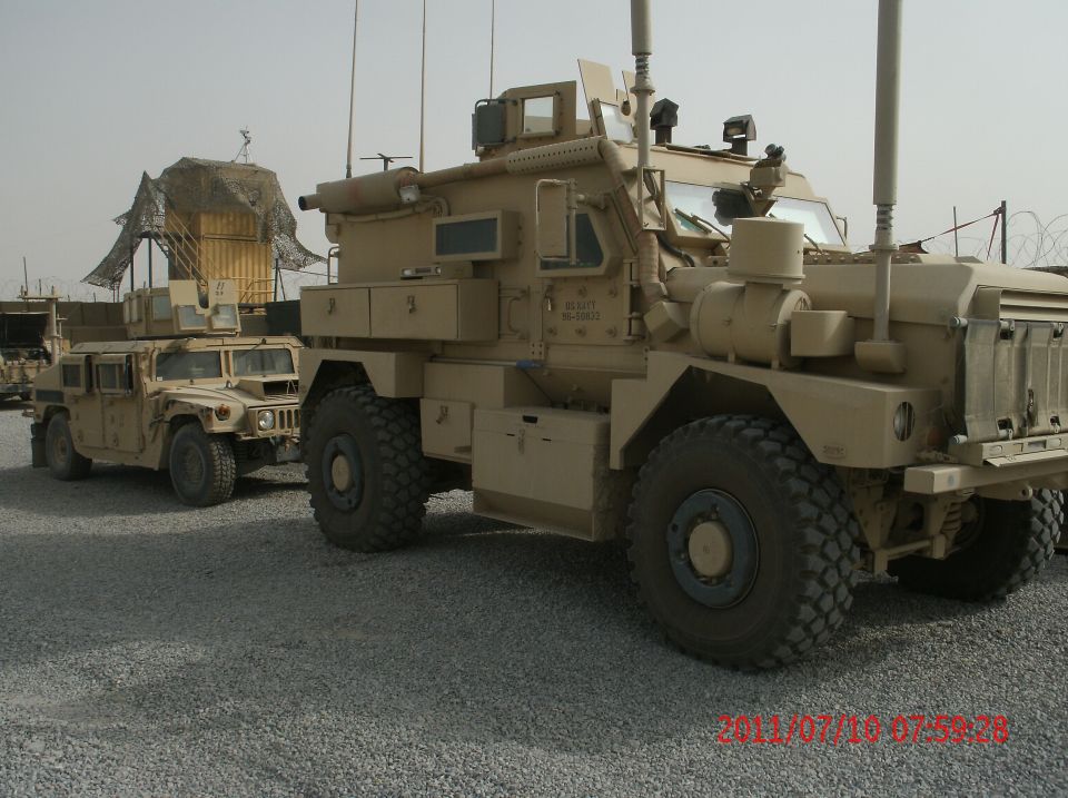 Two tan military trucks parked on a gravel lot. The truck in the foreground is larger and more heavily armored than the truck behind it